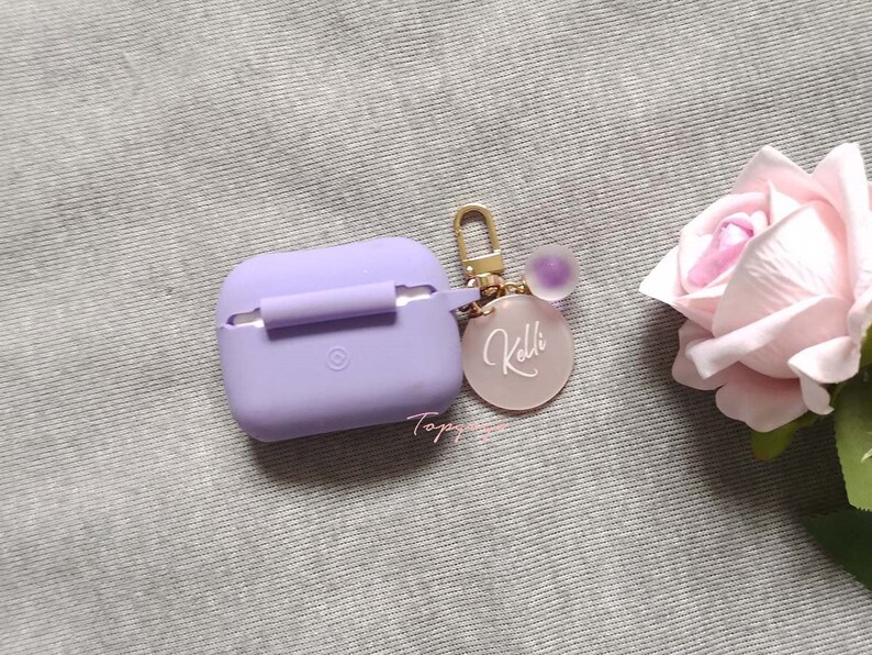 May include: A purple silicone case for wireless earbuds with a gold chain and a pink personalized charm that says "Kolbi".