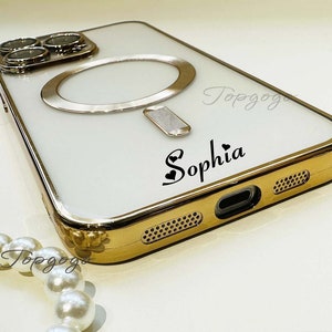 Puede incluir: Una funda blanca para iPhone con un borde dorado y un soporte para anillo. La funda tiene una inscripci&oacute;n personalizada que dice "Sophia" con un s&iacute;mbolo de coraz&oacute;n.