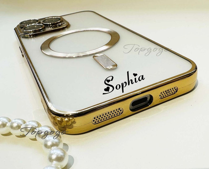 Puede incluir: Una funda blanca para iPhone con un borde dorado y un soporte para anillo. La funda tiene una inscripci&oacute;n personalizada que dice "Sophia" con un s&iacute;mbolo de coraz&oacute;n.