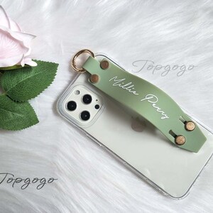 May include: A sage green phone grip with the name "Millie Perry" in white script. The grip is attached to a clear phone case with gold-colored rivets and a gold ring. A light pink rose and green leaves are in the background.