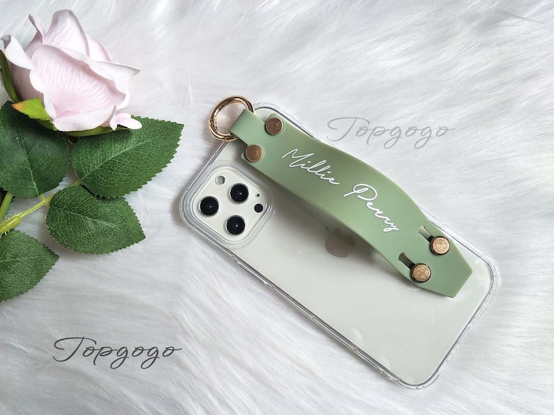 May include: A sage green phone grip with the name "Millie Perry" in white script. The grip is attached to a clear phone case with gold-colored rivets and a gold ring. A light pink rose and green leaves are in the background.