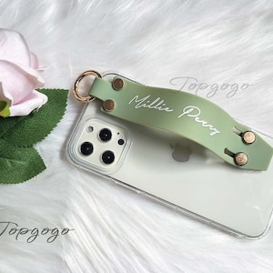 May include: A clear phone case with a light green strap featuring the name "Millie Perry" in white script. The strap has gold-colored rivets and a gold ring attachment. A pink rose and green leaves are in the background.