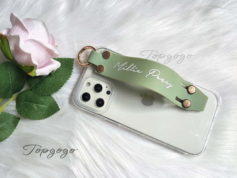 May include: A clear phone case with a light green strap featuring the name "Millie Perry" in white script. The strap has gold-colored rivets and a gold ring attachment. A pink rose and green leaves are in the background.