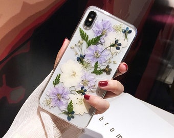 May include: A clear phone case featuring pressed flowers. The flowers are white, purple and blue. The case is designed for a smartphone with a dual camera lens.