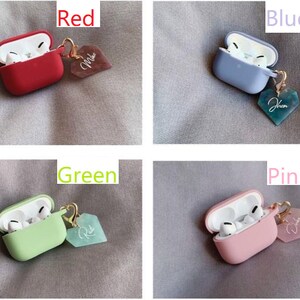 May include: Four Airpods cases with charms. The cases are red, blue-grey, green, and pink. Each case has a heart-shaped charm with a different color and inscription.