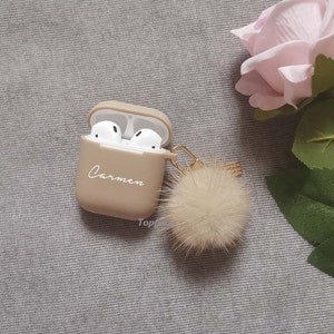 May include: A beige Airpods case with a fluffy pom-pom keychain. The case has the name "Carmen" written on it.