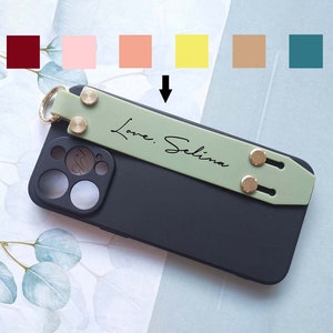May include: A black phone case with a green strap that says "Love. Selina". The strap has two gold metal accents. The phone case has a camera lens with multiple circles.