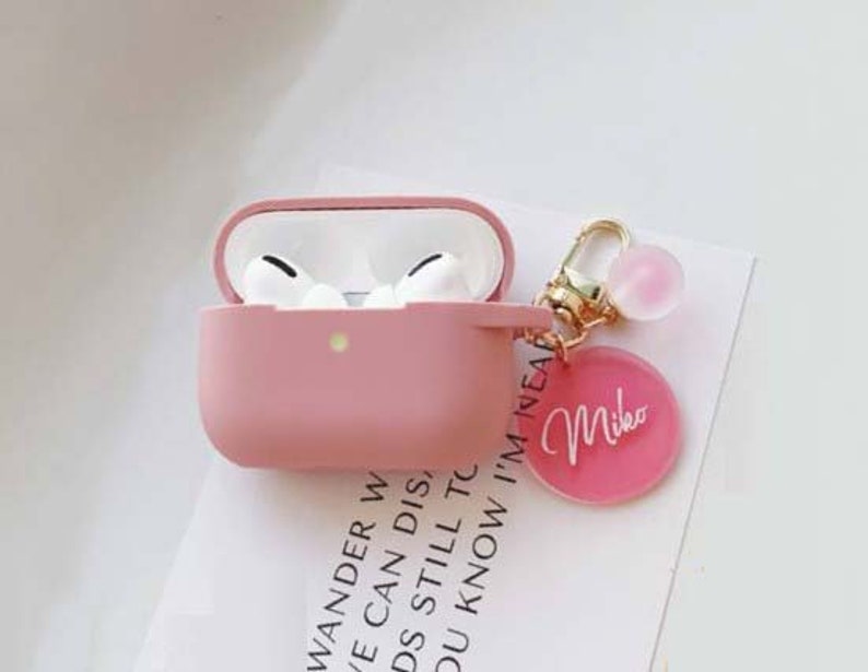 May include: Pink Airpods case with a gold keychain featuring a pink charm and a pink circle with the name "Mike" written in white.