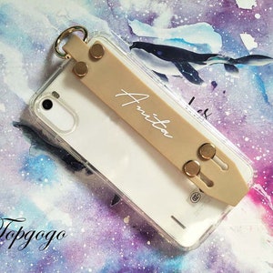 May include: A clear phone case with a beige phone grip attached. The grip has a gold ring and two gold snaps. The name "Anita" is written in white on the grip.