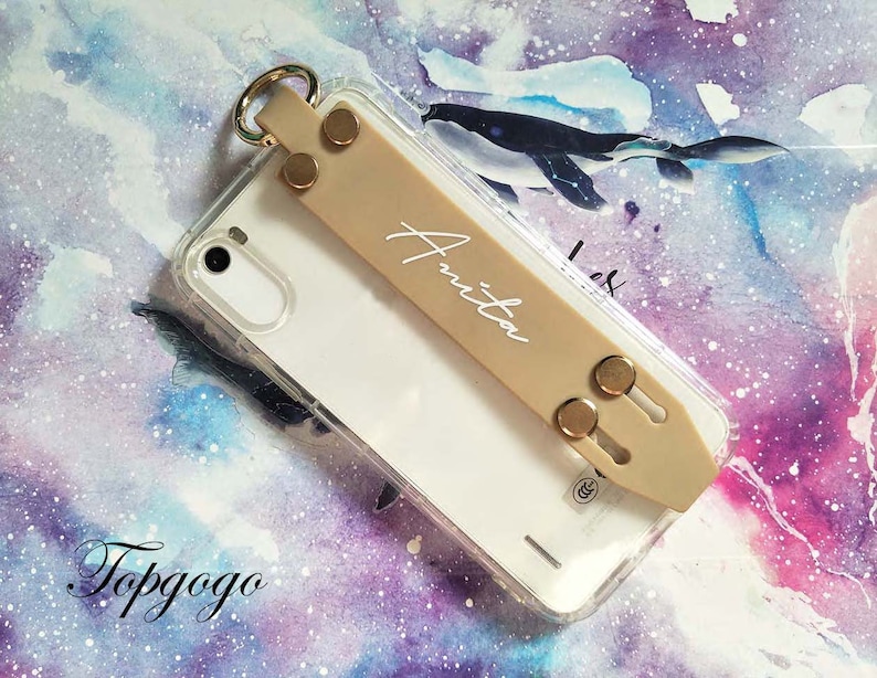 May include: A clear phone case with a beige phone grip attached. The grip has a gold ring and two gold snaps. The name "Anita" is written in white on the grip.