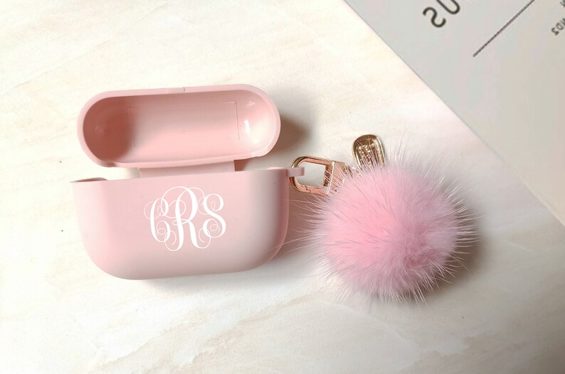 May include: A light pink AirPod case with a white monogram and a fluffy pink pom-pom keychain. The case is open, revealing the interior. The keychain has a gold-colored clasp.