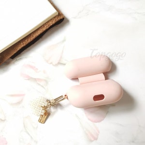 May include: A pink silicone case for wireless earbuds with a gold chain and a pearl charm attached. The case is open and the inside is visible.