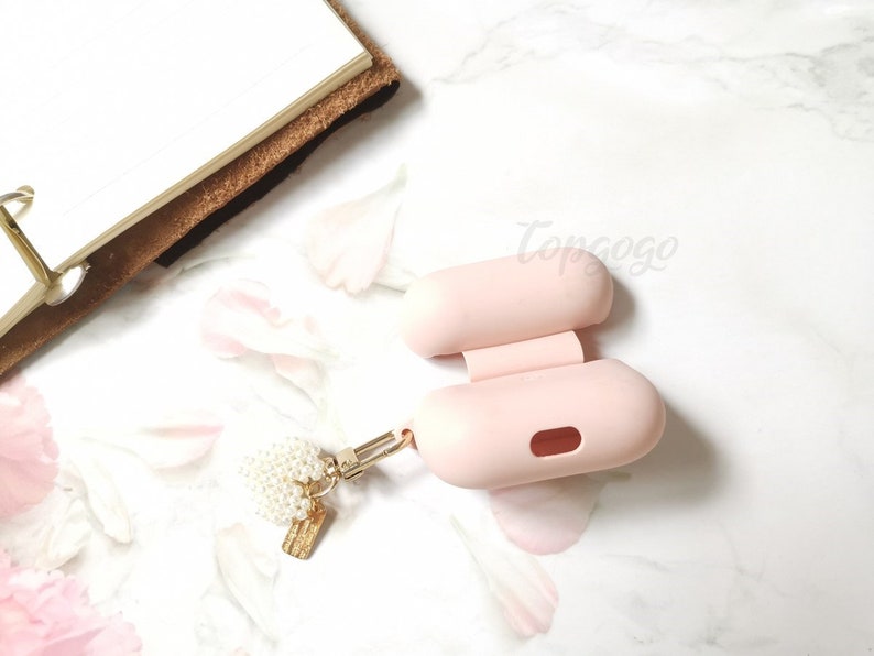 May include: A pink silicone case for wireless earbuds with a gold chain and a pearl charm attached. The case is open and the inside is visible.