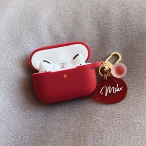 May include: Red Airpods case with a gold keychain featuring a red charm and a red oval tag with the name "Mika" written in white.