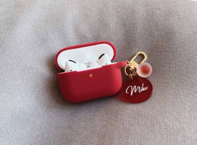 May include: Red Airpods case with a gold keychain featuring a red charm and a red oval tag with the name "Mika" written in white.