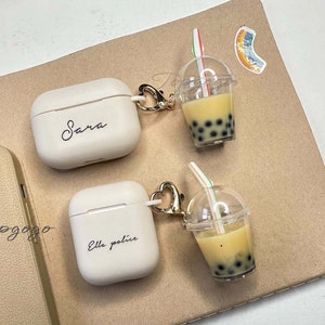 May include: Two white Airpod cases with personalized names "Sara" and "Ella police" attached to gold keychains with boba tea charms. The charms are clear plastic cups with a straw and brown boba pearls.