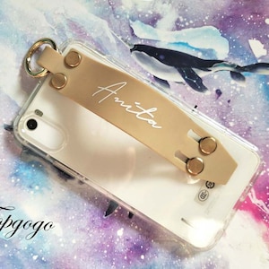 Può includere: Una custodia per telefono trasparente con un'impugnatura per telefono in pelle beige. L'impugnatura ha un anello dorato e due bottoni dorati. Il nome "Anita" è scritto in bianco sull'impugnatura. La custodia per telefono è su uno sfondo viola e rosa con un effetto acquerello.