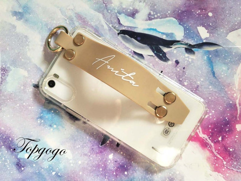 May include: A clear phone case with a beige leather phone grip. The grip has a gold ring and two gold buttons. The name "Anita" is written in white on the grip. The phone case is on a purple and pink background with a watercolour effect.