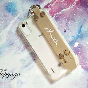 May include: A clear phone case with a beige strap that says "Anita" on it. The case has gold accents and is on a pink and purple watercolour background.