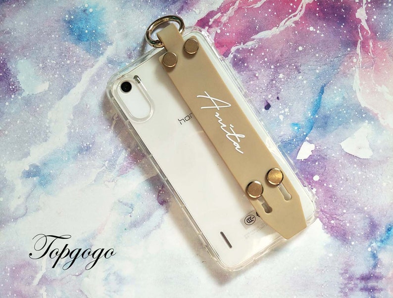 May include: A clear phone case with a beige strap that says "Anita" on it. The case has gold accents and is on a pink and purple watercolour background.