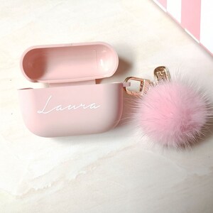 May include: A pink Airpods case with the name "Laura" written on it. The case is attached to a pink fluffy keychain.