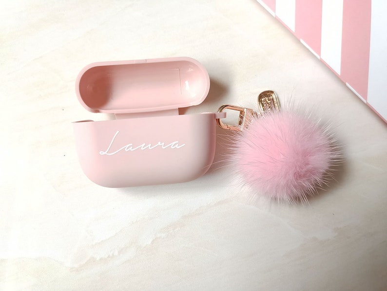May include: A pink Airpods case with the name "Laura" written on it. The case is attached to a pink fluffy keychain.