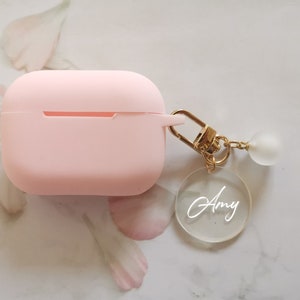 May include: A pink silicone case for wireless earbuds with a gold keychain attached. The keychain has a clear acrylic charm with the name "Amy" written in white.