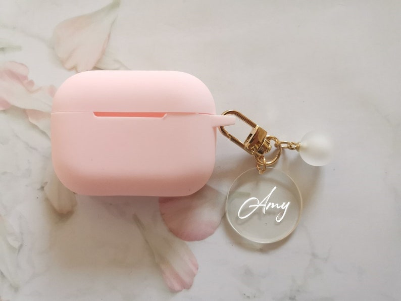 May include: A pink silicone case for wireless earbuds with a gold keychain attached. The keychain has a clear acrylic charm with the name "Amy" written in white.