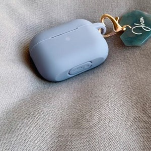 May include: A light blue silicone case for wireless earbuds with a gold keychain attached. The keychain has a green tag with the word "Jhen" written on it.