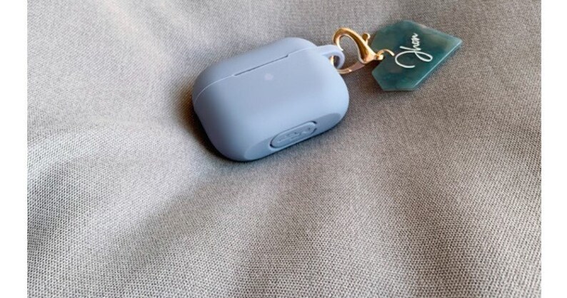 May include: A light blue silicone case for wireless earbuds with a gold keychain attached. The keychain has a green tag with the word "Jhen" written on it.