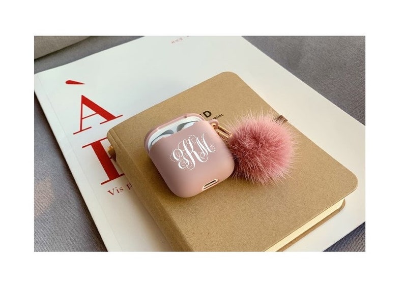 May include: A pink Airpods case with a white monogram "SRM" and a pink fluffy pom-pom keychain.
