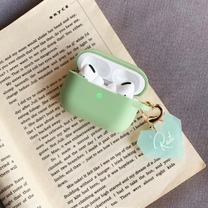 May include: A light green protective case for wireless earbuds with a gold keychain attached. The keychain has a light green tag with the word "Rait" written in cursive.