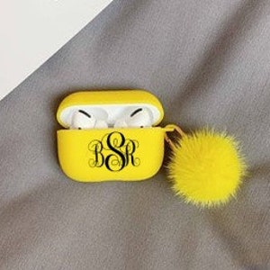 May include: A bright yellow AirPod case with a black monogram and a matching yellow pom-pom. The case is open, revealing white AirPod earbuds. The case is resting on a gray surface.