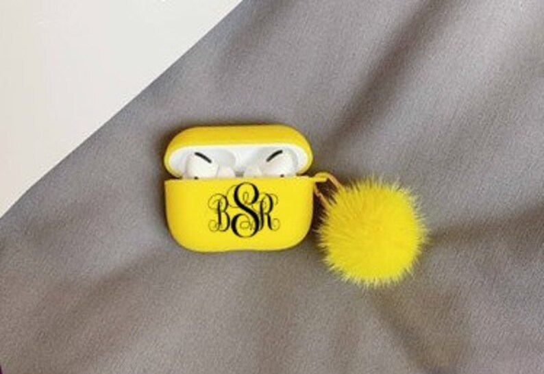 May include: A bright yellow AirPod case with a black monogram and a matching yellow pom-pom. The case is open, revealing white AirPod earbuds. The case is resting on a gray surface.