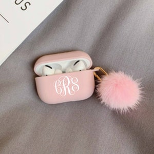 May include: A light pink AirPods Pro case with a white monogram and a fluffy pink pom-pom keychain. The case is open, revealing the white earbuds inside. The case is resting on a gray surface.