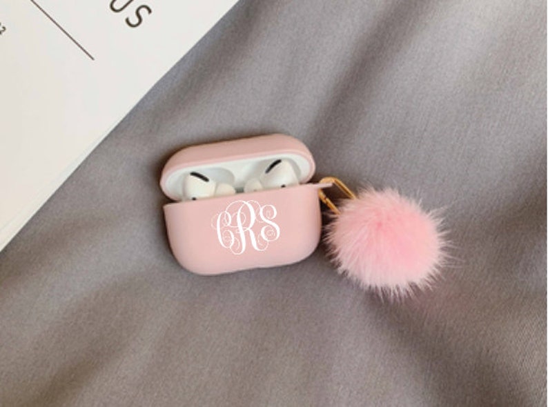 May include: A light pink AirPods Pro case with a white monogram and a fluffy pink pom-pom keychain. The case is open, revealing the white earbuds inside. The case is resting on a gray surface.