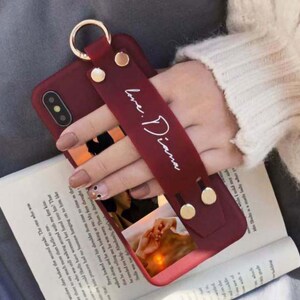 May include: A red phone case with a gold ring and a red strap that says "Love: Diana". The case has a photo of a couple holding hands in the background.