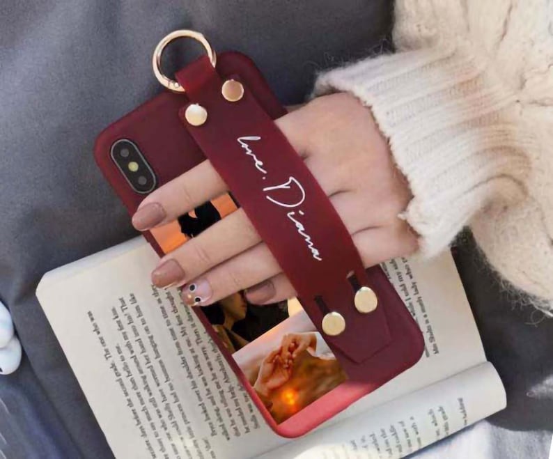 May include: A red phone case with a gold ring and a red strap that says "Love: Diana". The case has a photo of a couple holding hands in the background.