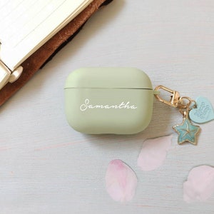May include: A light green Airpods case with the name Samantha written on it. The case has a gold chain attached to it with a blue starfish charm and a heart charm that says "Blessed" on it.