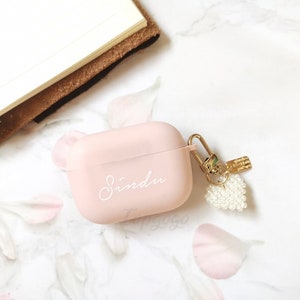 May include: A pink Airpods case with a gold keychain and a pearl heart charm. The case has the name "Sindu" written on it in white.