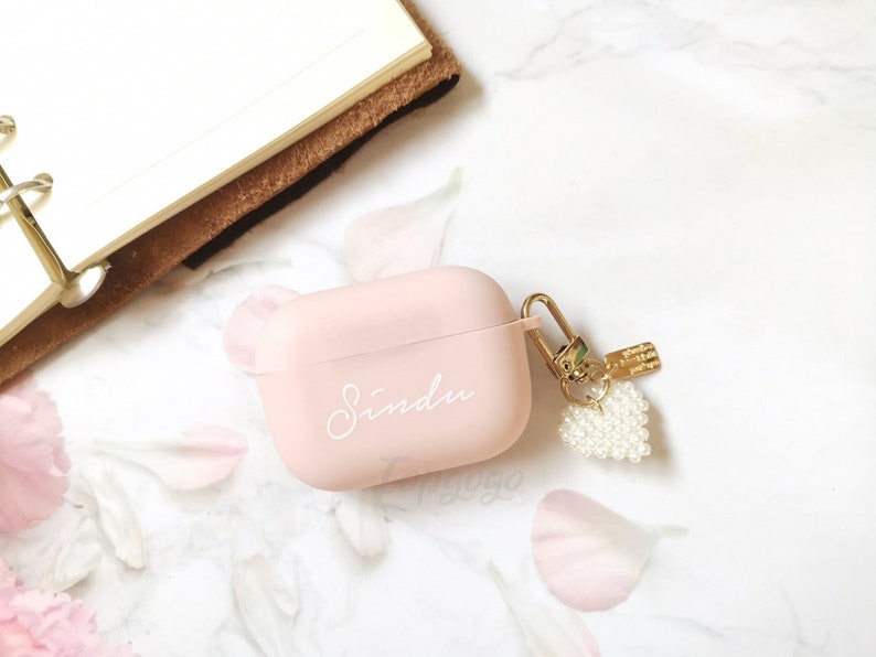 May include: A pink Airpods case with a gold keychain and a pearl heart charm. The case has the name "Sindu" written on it in white.