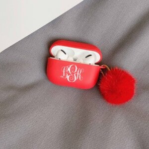 May include: Red AirPods case with a white monogram and a matching red pom-pom keychain. The case is open, revealing white earbuds. The background is a gray fabric.