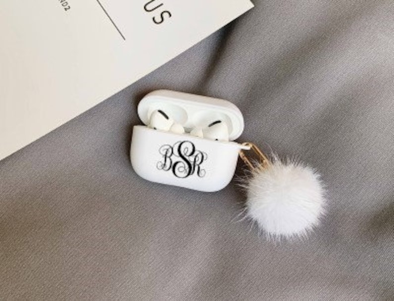 May include: White AirPods Pro case with a black monogram and a fluffy white pom-pom keychain. The case is open, revealing the earbuds. The background is a light gray fabric.