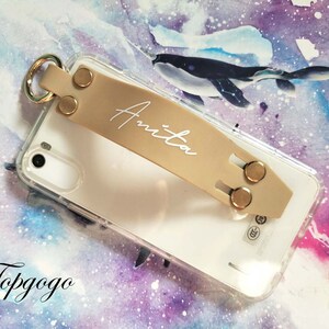 May include: A beige phone case with a clear back and a gold ring attached. The case has a beige leather strap with a gold buckle and the name "Anita" written in white on the strap.