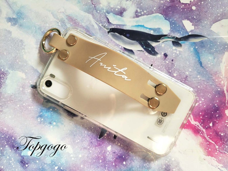 May include: A beige phone case with a clear back and a gold ring attached. The case has a beige leather strap with a gold buckle and the name "Anita" written in white on the strap.