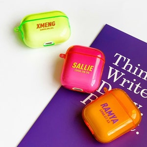 K&ouml;nnte beinhalten: Drei transparente AirPod-H&uuml;llen aus Kunststoff mit personalisierten Namen und Daten. Die H&uuml;llen sind gr&uuml;n, rosa und orange. Die H&uuml;llen befinden sich auf einer violetten Oberfl&auml;che mit dem aufgedruckten Text "Think Write Read".