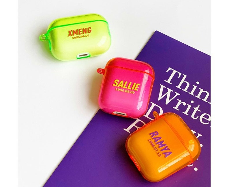 K&ouml;nnte beinhalten: Drei transparente AirPod-H&uuml;llen aus Kunststoff mit personalisierten Namen und Daten. Die H&uuml;llen sind gr&uuml;n, rosa und orange. Die H&uuml;llen befinden sich auf einer violetten Oberfl&auml;che mit dem aufgedruckten Text "Think Write Read".