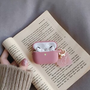 May include: A pink protective case for wireless earbuds with a gold charm that says "Dube". The case is open and the earbuds are visible inside.