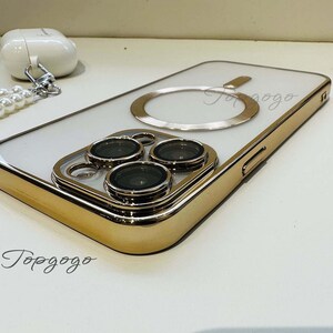 Puede incluir: Una funda blanca para iPhone con un borde dorado y un anillo dorado en la parte posterior. La funda tiene tres lentes de c&aacute;mara en la parte posterior.