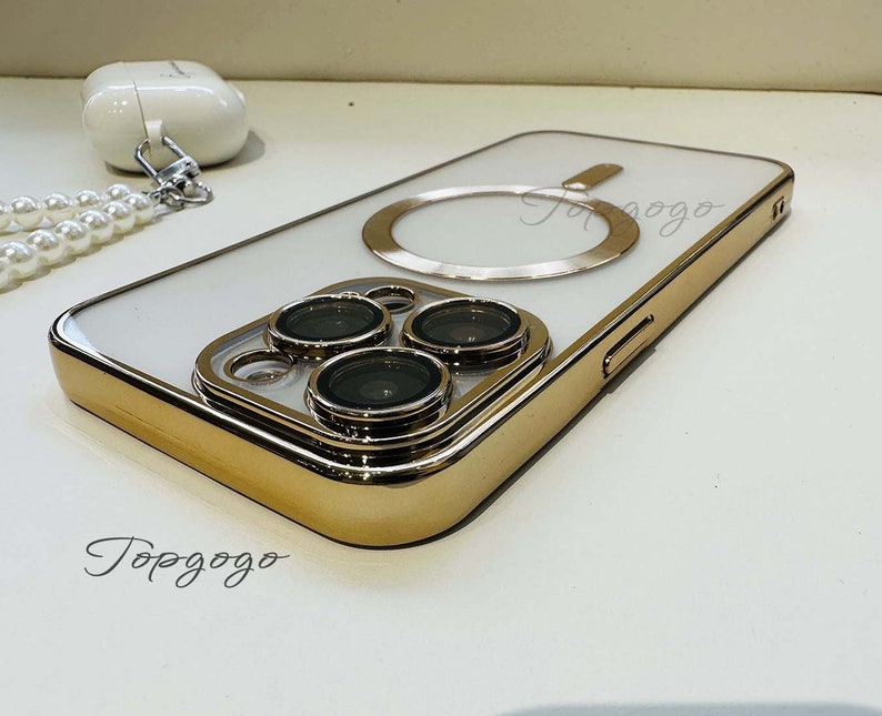 Puede incluir: Una funda blanca para iPhone con un borde dorado y un anillo dorado en la parte posterior. La funda tiene tres lentes de c&aacute;mara en la parte posterior.